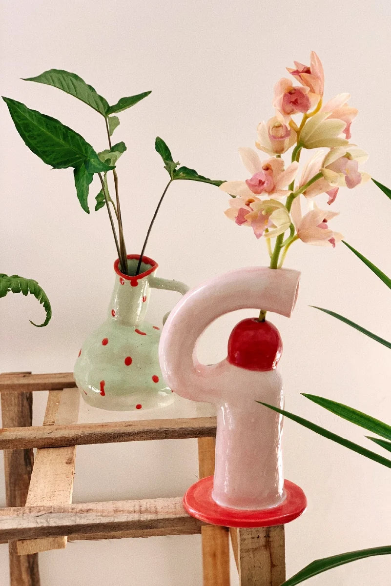 Decorative vase shaped like a letter 'F' with pink flowers on a wooden stool against a white wall.